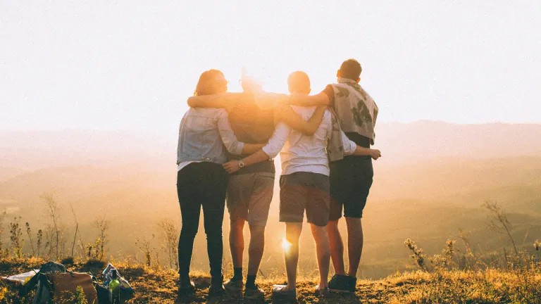 Four people with arms over each other's shoulders facing away with sun shining